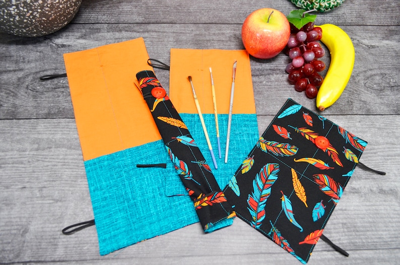 May include: Two fabric pencil cases with a black background and colourful feather pattern. The cases are rolled up and have a button closure. One case is open and has four paintbrushes inside. The cases are on a wooden surface with a banana, grapes, and an apple.