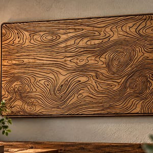 May include: A rectangular wooden wall plaque featuring a dark, laser-engraved topographic map pattern with intricate, flowing contour lines that mimic natural wood grain patterns across the light brown surface of the board.
