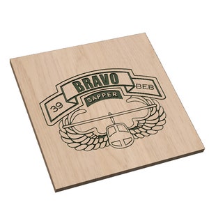 Puede incluir: Una placa de madera con un diseño grabado en verde y negro de una insignia militar. La insignia presenta un helicóptero, alas y el texto "BRAVO SAPPER 39 BEB".