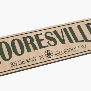 Puede incluir: Un letrero de madera con el texto "MOORESVILLE" en letras verdes y las coordenadas "35.58486° N -80.81007° W" debajo. Una rosa de los vientos se encuentra entre el texto y las coordenadas.