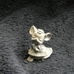May include: A small, silver-colored mouse figurine with large ears and a smiling face. The mouse is seated on a decorative base, possibly a nut or shell. The figurine has a vintage or antique appearance.