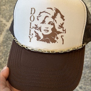 May include: A brown and white trucker hat with a brown silhouette of Dolly Parton and the word "DOLLY" printed on the front. The hat has a gold chain trim around the brim.