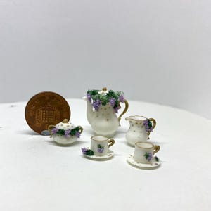 Puede incluir: Juego de té de porcelana en miniatura con detalles florales. El juego incluye una tetera, una jarra de crema, un azucarero y dos tazas de té con platillos. Cada pieza está decorada con flores moradas y ribetes dorados. Se incluye una moneda para la escala.
