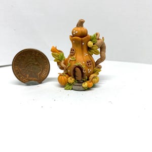 May include: A miniature pumpkin-shaped teapot house sculpture featuring a small arched door, windows, and vines. The orange and brown clay piece is displayed next to a copper penny for scale, highlighting its small size and detailed texture.