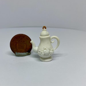 May include: A small, off-white ceramic teapot with a decorative floral design. The teapot has a curved handle and spout, and a small loop on the lid. A penny is next to the teapot for size comparison.