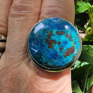 May include: A round ring featuring a large, polished cabochon stone with a vibrant mix of deep blue, turquoise, and dark brown matrix patterns. The stone is set in a simple, smooth silver-toned metal bezel, creating a bold and eye-catching piece.