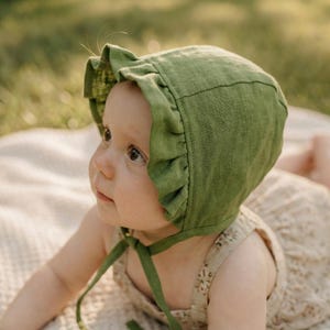 May include: A green linen baby bonnet with a frilled edge and tie fastening. The bonnet is a soft, muted green colour. The baby is wearing a floral print romper.