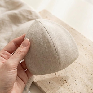 May include: A beige, triangular-shaped fabric item, possibly a bra cup or similar garment component, is held by a hand. The fabric has a textured appearance and a seam running along the top edge. The background is a light-colored surface.