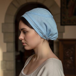 May include: A light blue linen head covering, likely a bonnet or cap, is worn. The headwear is soft and draped, with a structured top and a band around the head. The style suggests historical or vintage fashion.
