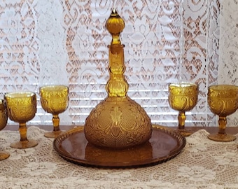 May include: Amber-colored glass drinkware set. Includes a decanter with a stopper, a round tray, and six stemmed glasses. The glassware features an embossed floral design. The set is arranged on a lace tablecloth.