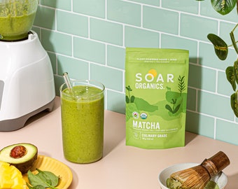 May include: A green smoothie in a glass with a straw, a bag of Soar Organics Culinary Grade Matcha green tea powder, a bowl of matcha powder with a whisk, and a sliced avocado and mango on a yellow plate.