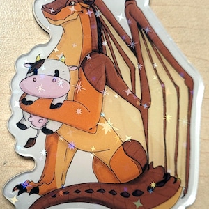 May include: A sticker featuring a cartoon dragon with brown and orange scales, holding a plush cow. The dragon has large wings and is sitting down. The sticker has a holographic effect with star-shaped sparkles.