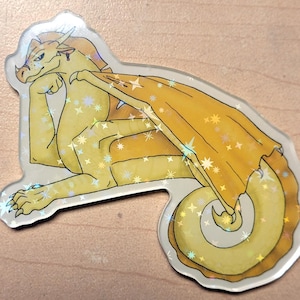 May include: A yellow dragon sticker with iridescent accents. The dragon is in a relaxed pose, with one arm supporting its head. The wings and body are detailed with star-shaped sparkles. The sticker has a clear, glossy border.