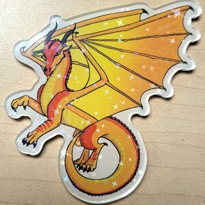 May include: A holographic sticker featuring a stylized dragon with orange, yellow, and red scales. The dragon has large wings and is posed with its tail curled. The sticker has a clear border and a sparkly, iridescent finish.