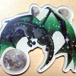 May include: A holographic sticker featuring a dragon with green wings and a starry night design. The dragon is surrounded by three moons and various stars. The sticker has a glossy finish.