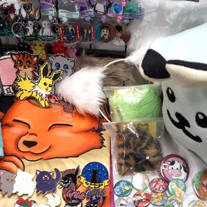 May include: A collection of handmade items including plush toys, embroidered patches, pins, and keychains. The items feature various cartoon characters and designs, including a blue plush toy with black ears and a white face, a yellow and orange fox plush, and a blue and pink Pokemon character.