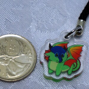 May include: A clear plastic charm with a cartoon dragon design. The dragon is green with blue and red accents. The charm is attached to a black cord.