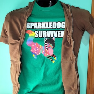 Puede incluir: Una chaqueta marrón con cremallera y una camiseta verde debajo. La camiseta presenta un zorro de dibujos animados colorido con el texto "Sparkledog Survivor".