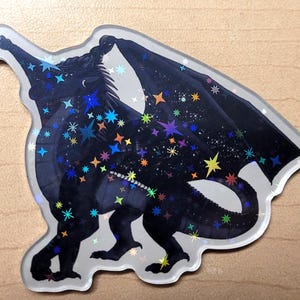 May include: A holographic sticker featuring a black dragon with outstretched wings. The dragon is covered in colorful stars and a crescent moon. The sticker has a white border and is on a light brown surface.