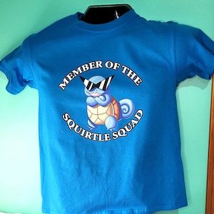 Puede incluir: Una camiseta azul brillante con un dibujo de Squirtle con gafas de sol y el texto "MEMBER OF THE SQUIRTLE SQUAD". El diseño es circular. La camiseta se muestra en una cabeza de maniquí negra.