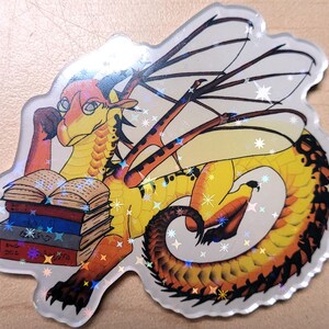 May include: A holographic sticker featuring a yellow and orange dragon reading books. The dragon has large wings and is resting its head on its arm. The sticker has a sparkly, iridescent finish and is on a light brown surface.