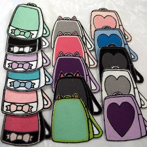 May include: A collection of 12 colourful felt backpack patches with various designs, including hearts, bows, and cat ears. The patches are all different colours, including pink, blue, green, purple, and black.