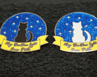 May include: Two enamel pins with a blue glitter background and a white ribbon with the text "May StarClan light your path". One pin features a black cat silhouette and the other features a white cat silhouette.