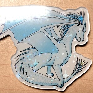 May include: A holographic sticker featuring a light blue dragon with outstretched wings and a long tail. The dragon is detailed with blue accents and star-shaped sparkles. The sticker has a clear, glossy finish.