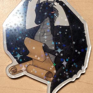 May include: A holographic sticker featuring a black dragon with outstretched wings, holding a scroll. The dragon is detailed with gray scales and adorned with star-shaped accents. The sticker has a clear, irregular border.