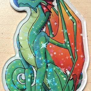 May include: A holographic sticker featuring a cartoon dragon in shades of green, blue, and orange. The dragon is sitting with its wings spread, and the sticker has a clear, glossy finish with star-shaped accents.
