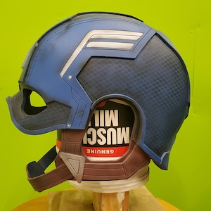 May include: A blue and white Captain America helmet with a textured surface. The helmet is on a brown base with a red and white label that says "GENUINE MUSC"