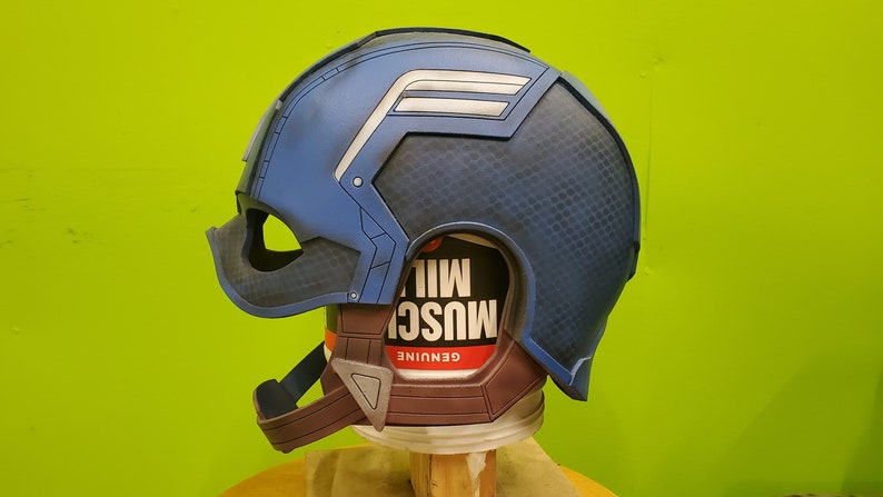 May include: A blue and white Captain America helmet with a textured surface. The helmet is on a brown base with a red and white label that says "GENUINE MUSC"