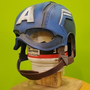 May include: A blue and white Captain America helmet with a brown chin strap. The helmet is made of plastic and has a textured surface. The helmet is on a wooden stand.