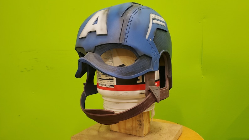 May include: A blue and white Captain America helmet with a brown chin strap. The helmet is made of plastic and has a textured surface. The helmet is on a wooden stand.