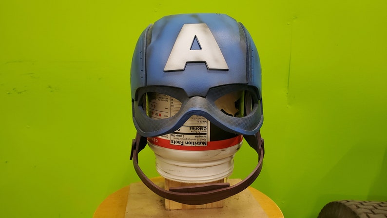 May include: A blue and white Captain America helmet with a red and white strap. The helmet is made of plastic and has a distressed look.