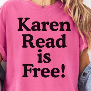 Puede incluir: Una camiseta rosa brillante con el texto negro "Karen Read is Free!". La camiseta tiene mangas enrolladas y se combina con una chaqueta vaquera. El estilo es informal y cómodo.