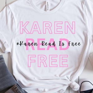 Puede incluir: Camiseta blanca con el texto rosa "KAREN READ IS FREE" y el hashtag "#Karen Read Is Free". La camiseta está combinada con vaqueros grises, un par de zapatillas de deporte azul marino, un libro y una taza de café.