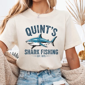 May include: A cream-coloured t-shirt featuring a blue and white shark design. The text on the shirt says "QUINT'S SHARK FISHING EST. 1975". The t-shirt is short-sleeved with a crew neck.