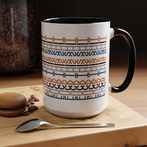 Puede incluir: Taza de café de cerámica blanca con asa y borde negros. La taza presenta un patrón geométrico repetitivo en tonos marrón, gris y azul. La taza está sobre una superficie de madera con una cuchara y un macaron.