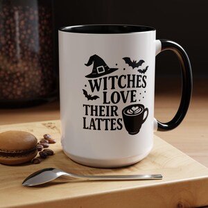 May include: White ceramic mug with a black interior and handle. The mug features the text "Witches Love Their Lattes" with witch hat, bat, and coffee cup illustrations. A spoon and a macaron are on a wooden surface.