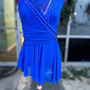 May include: A vibrant blue dance dress with sparkling embellishments. The sleeveless dress features a draped bodice and a flared skirt, adorned with scattered rhinestones. The dress is displayed on a red dress form, showcasing its design and details.