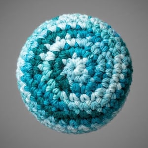 May include: A round, crocheted ball in shades of teal, turquoise, and white. The yarn is tightly woven, creating a textured surface. The ball is a solid color, with no visible seams or embellishments. The background is a solid gray.