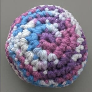 May include: A round, hand-crocheted sphere featuring a variegated yarn pattern in shades of purple, light blue, and white. The textured surface is created with dense, spiraling crochet stitches that form a consistent, multi-colored circular design.