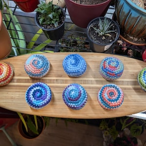 May include: Eight round, crocheted spheres featuring various multi-colored spiral patterns in shades of blue, green, orange, and purple. These textured, yarn-based items are arranged in two rows on an elongated, oval-shaped wooden serving board.