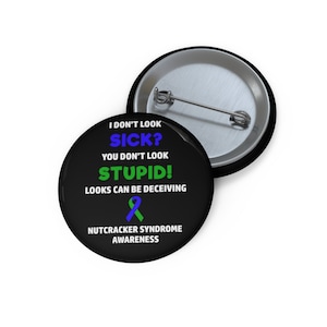 May include: A black button with the text "I DON'T LOOK SICK? YOU DON'T LOOK STUPID! LOOKS CAN BE DECEIVING" in white, blue, and green. The button also features a blue and green ribbon and the words "NUTCRACKER SYNDROME AWARENESS". A second button is visible.