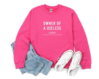 Peut inclure: Sweat-shirt rose avec le texte "OWNER OF A USELESS colon" en blanc. Le sweat-shirt est plié et posé sur un jean bleu et des baskets blanches.
