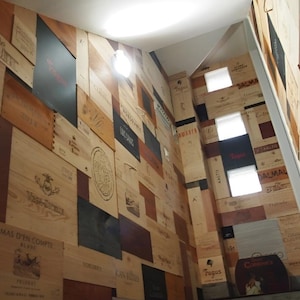 Può includere: Un'installazione a parete composta da numerosi pannelli rettangolari in legno provenienti da casse di vino, disposti a mosaico. I pannelli presentano varie tonalità di legno naturale, accenti neri e loghi, testi e illustrazioni stampate.