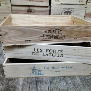 May include: Three wooden wine crates stacked on top of each other. The top crate has the text "CHATEAU DUHART-MILON 2020". The middle crate reads "LES FORTS DE LATOUR 2017". The bottom crate says "PAGODES DE COS 2021".