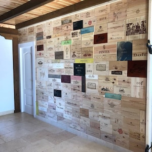 Pode incluir: Uma parede decorada com numerosos painéis de caixas de vinho de madeira. Os painéis são de vários tamanhos e apresentam textos e logotipos impressos em diferentes cores. A parede faz parte de uma sala com uma porta branca e um chão claro.