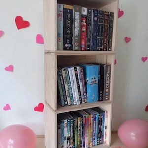 Może przedstawiać: Trzypoziomowy, jasny drewniany regał wypełniony płytami DVD i zestawami seriali telewizyjnych. Jednostka jest ozdobiona różowymi i czerwonymi wycięciami w kształcie serca oraz dwoma różowymi balonami. Widoczne tytuły to "Deadwood" i "Game of Thrones."